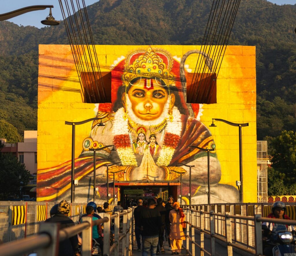 Vibrant Hanuman mural on a bridge in Rishikesh, Uttarakhand, with people and mountain backdrop.